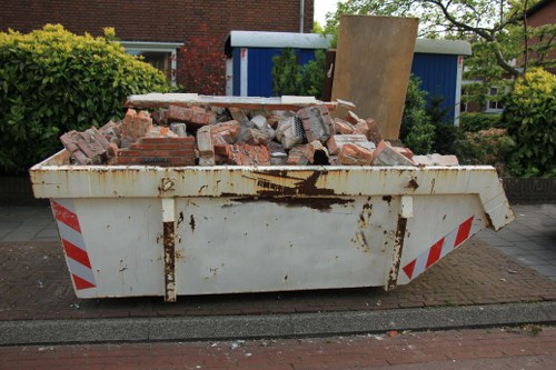 A large white skip placed on a paved street surface in an urban residential area, filled with a mixed pile of red brick debris and broken concrete blocks. The skip shows signs of rust and dirt on its exterior, with red and white reflective hazard stripes on the side panels for safety. In the background, there are green bushes and trees, alongside a blue shed or container, and brick buildings with windows, indicating a suburban setting. The scene suggests that a rubbish removal or demolition cleanup operation, managed by Commercial Waste Manor Park, is taking place in the local area of Manor Park, within the postal code vicinity. The image captures the typical environment for waste disposal services, with an emphasis on the collection of construction or building debris, suitable for clients seeking professional rubbish removal.