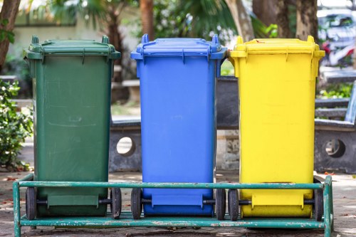 In an outdoor urban setting, three wheeled bins are positioned side by side on a paved surface, each with a distinct color: green on the left, blue in the middle, and yellow on the right. The green bin exhibits a smooth, matte plastic surface with slight scratches, while the blue bin appears similarly made of plastic with a shiny finish, reflecting ambient light. The yellow bin, also of plastic, features a matte texture with visible ridges along its lid. All three bins are mounted on black rubber wheels, with the green and blue bins slightly leaning forward and the yellow bin standing more upright. Behind the bins, there are benches with wooden planks and metal frames, and in the background, there are trees with green foliage and parked cars, indicating a communal outdoor area such as a park or street frontage. The scene suggests waste management activity typical of local rubbish removal services offered by Commercial Waste Manor Park, with natural daylight illuminating the area clearly, emphasizing the different colors and textures of the waste containers.