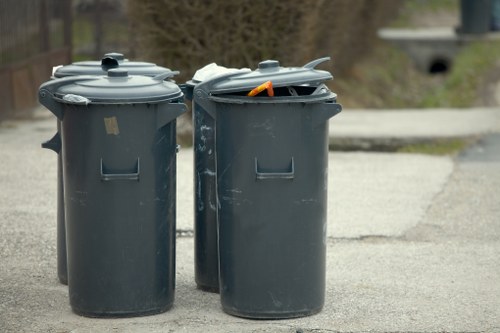 Two large, dark grey plastic wheelie bins with fitted lids are positioned on a paved driveway in an outdoor setting. The bin on the left has a closed lid with a small, rectangular sticker or label on top. The bin on the right has an open lid, revealing a partially visible orange-handled tool or object inside. Behind the bins, the background features a blurred view of a grassy area, some trees, and possibly a low brick or stone wall, indicating a residential or suburban environment. The surface of the bins shows minor scuff marks, and they are situated on a concrete or tarmac surface, typical for waste collection points. The overall scene suggests a routine rubbish collection spot, consistent with services offered by Commercial Waste Manor Park in the area near postcode inferred for the town in east London.