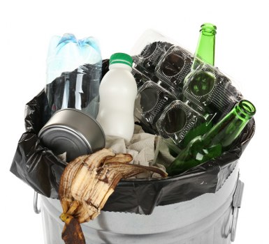The image shows a metal waste bin lined with a black plastic bag, filled with various types of discarded rubbish. Inside the bin, there is a crumpled plastic bottle with a light blue label, a white plastic milk bottle with a green cap, a silver metal can, and several dark glass bottles, some with green coloration, all scattered and partly visible. Additionally, there are a few pieces of torn cardboard or paper and other mixed waste. The bin appears to be placed outdoors, possibly on a driveway or paved area, with the background not visible. The scene depicts typical household rubbish awaiting disposal, relevant to rubbish removal services such as those offered by Commercial Waste Manor Park, servicing local areas including Manor Park and surrounding districts in east London.