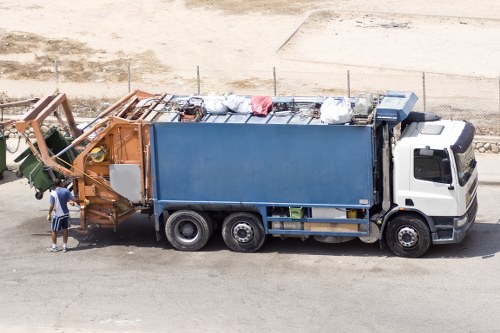 A large waste collection vehicle parked along a street in a suburban area, with a white cab and a blue container body used for rubbish collection. The vehicle is positioned on a paved driveway or road, with the front facing to the right. Visible on the left side, a worker wearing a high-visibility vest, dark trousers, and a cap is standing near the rear of the vehicle, appearing to either load or inspect the collected waste. The waste in the container includes various materials covered with protective plastic sheets, with some loose rubbish visible on top. In the background, there is a sandy or gravelly surface, possibly part of a construction or storage yard, bordered by fencing. The scene suggests a commercial waste removal operation typical of rubbish collection services in the Manor Park area, with the vehicle ready for or just having completed waste collection tasks.