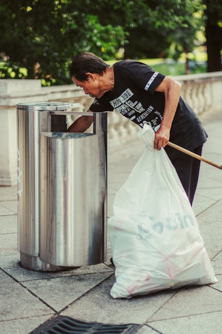 A middle-aged man with dark hair wearing a black T-shirt and dark pants is seen using a metal waste bin on a paved sidewalk. He is bending slightly forward, holding a long-handled tool in his left han
