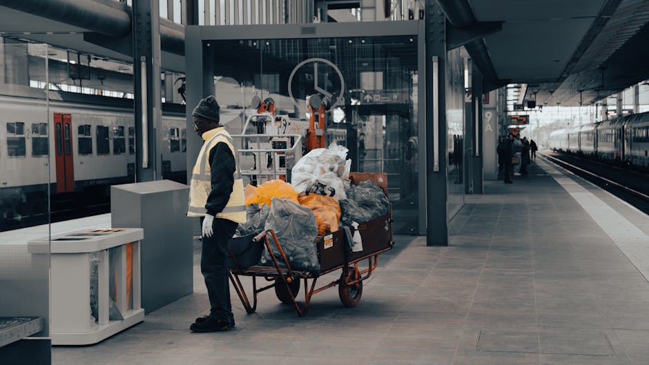 A man wearing a dark jacket, black trousers, white gloves, and a grey knit beanie stands on a modern train station platform. He is positioned next to a large, orange, metal trolley filled with various