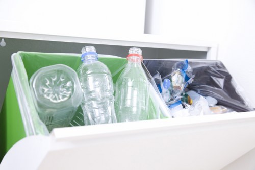 A close-up image of a white waste collection container with two transparent plastic bottles placed inside a green compartment on the left side and a black garbage bag filled with assorted waste items in a separate black section on the right. The bottles, one with a blue cap and the other with a red cap, appear to be discarded beverage containers. The container is situated on a smooth, light-colored surface, likely within an indoor or outdoor setting associated with waste disposal or rubbish collection services. The scene reflects typical materials handled during rubbish removal, with clear emphasis on plastic waste, indicative of the type of refuse managed by Commercial Waste Manor Park as part of their sustainable commercial waste solutions in the area.