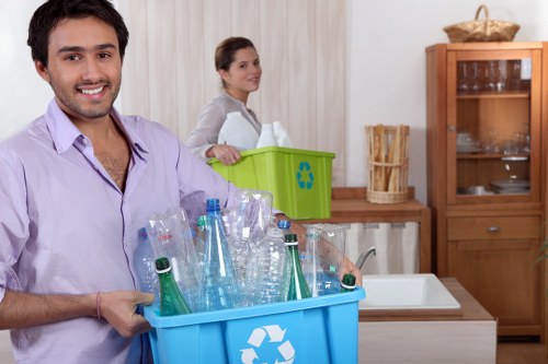 A man with a cheerful expression holds a bright blue recycling bin filled with clear plastic bottles, including some with green and blue tint, in a kitchen setting. Behind him, a woman stands with a neutral expression next to a green recycling bin that features a recycling symbol, positioned on a wooden countertop next to a fabric basket and a wooden cabinet with glass doors. The kitchen has light-colored wall panels, a white sink, and various kitchen essentials, indicating a domestic environment. The scene highlights waste collection and recycling activities within a home in Manor Park, suggesting a focus on sustainable rubbish management services. The natural lighting emphasizes the clarity of the bottles and the textures of the bin and kitchen surfaces, supporting the context of rubbish and waste disposal for local clients such as Commercial Waste Manor Park.