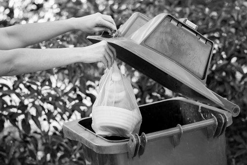 A person placing a white plastic bag filled with household rubbish into a dark-colored, open waste bin situated outdoors on a paved surface, with a background of leafy shrubs or hedges. The lid of the bin is propped open, revealing a clean, textured interior. The scene suggests a routine rubbish disposal activity, and the setting appears to be in a residential or commercial area within Manor Park, aligning with services offered by Commercial Waste Manor Park for waste collection and rubbish removal. The overall environment is well-lit, with natural daylight illuminating the scene, emphasizing the practical, everyday nature of waste management tasks.