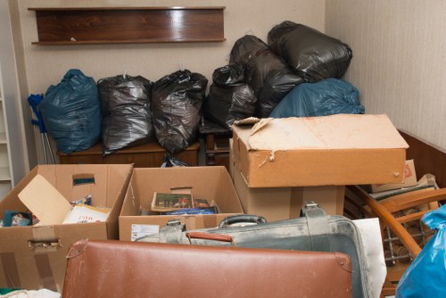 A cluttered indoor storage area or garage containing multiple black rubbish bags arranged in a row along a wooden shelf, with their tops twisted and secured, and several blue bags positioned nearby. In the foreground, open cardboard boxes filled with miscellaneous items, including papers and small objects, are stacked on top of one another. A large, grey plastic vacuum cleaner or similar equipment lies on the floor, partly covered by the boxes. To the right, a small wooden stool or step ladder is visible, with a piece of brown furniture or partition at the lower part of the image. The background features a beige wall with a wooden shelf mounted above the bags. The scene appears to be an organized but cluttered area awaiting rubbish removal or clearing, typical of a domestic property in Manor Park, with the company's name, Commercial Waste Manor Park, associated with rubbish collection services.